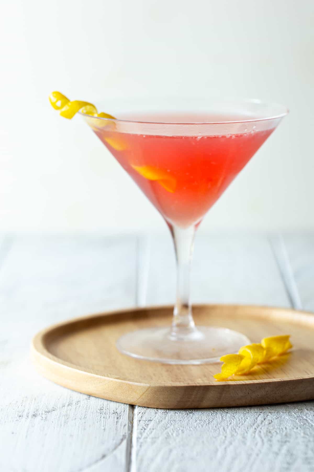 single martini glass filled with pink drink on wooden tray with lemon twist