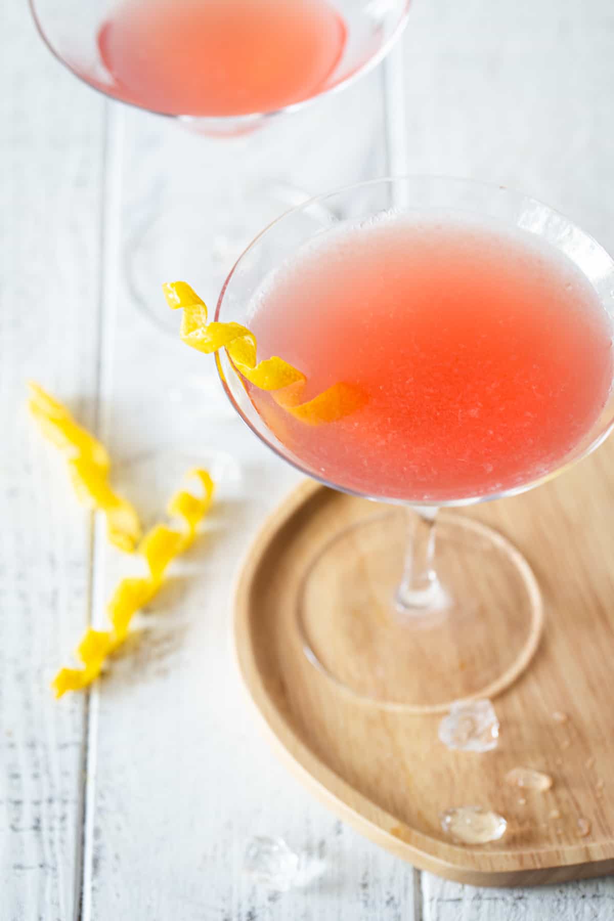 ¾ angle of martini glass on wooden tray willed with pink drink and garnished with lemon twist