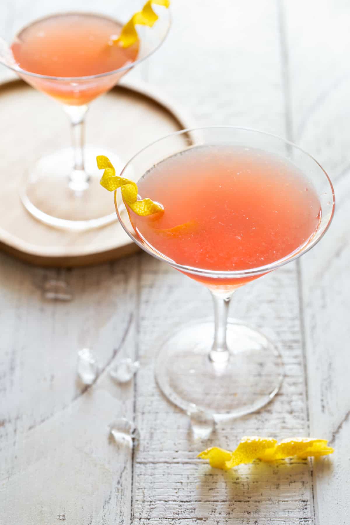 tall martini glass filled with cosmo on a white background with second glass in background on a wooden plate and a lemon twist.