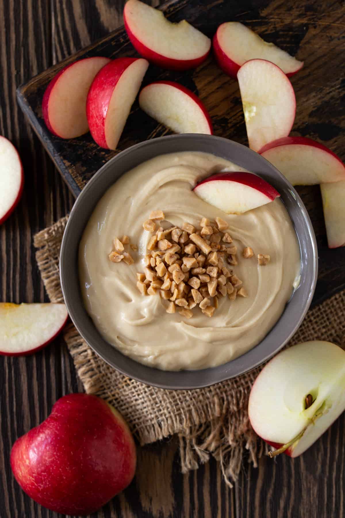 Bowl of dip topped with toffee bits and sliced apples on board.