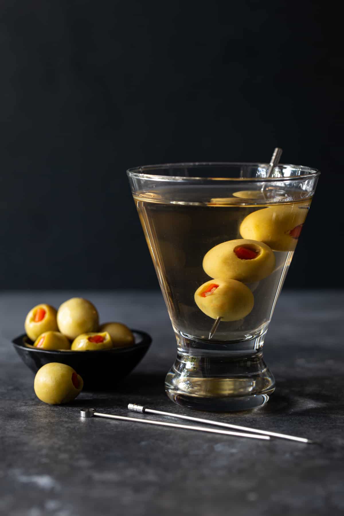 bowl of olives with metal skewers on board and filled martini glass with olives in it
