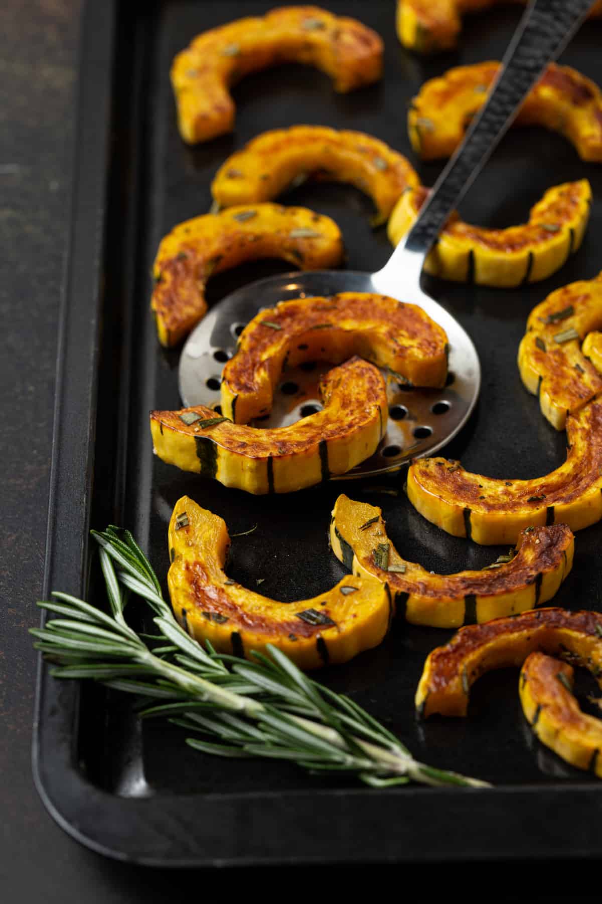 Roasted squash pieces on a dark sheet pan with silver lifting utensil and fresh rosemary. 