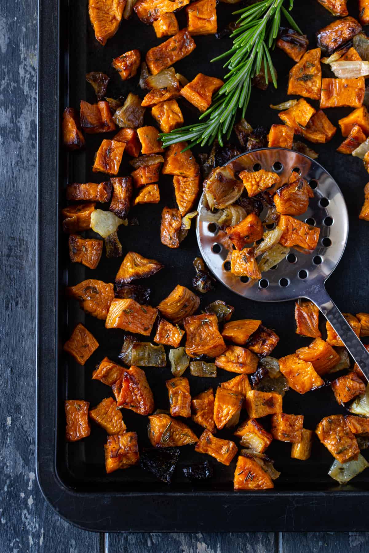 Dark sheet pan filled with roasted sweet potatoes and onions. Silver spoon lifting potatoes from pan. 