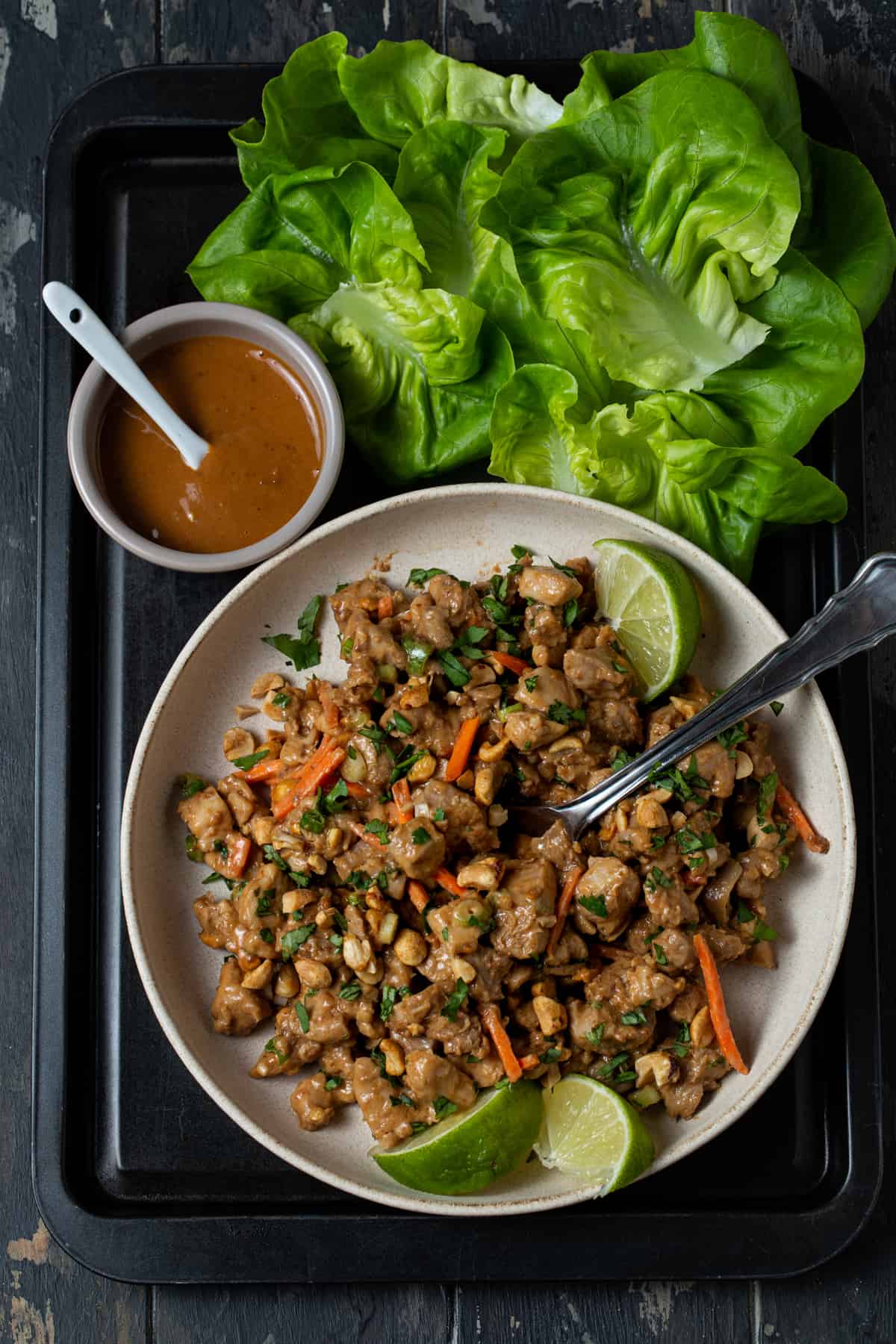 chopped chicken filling for lettuce wraps , bowl of peanut sauce and lettuce cups on black tray.