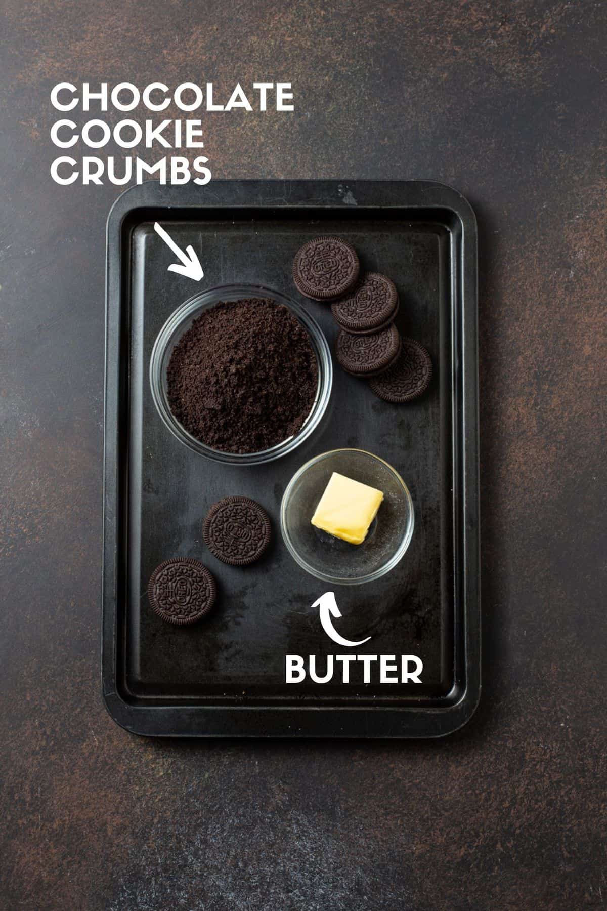 Dark sheet pan with chocolate cookies, bowl of cookie crumbs and butter. 