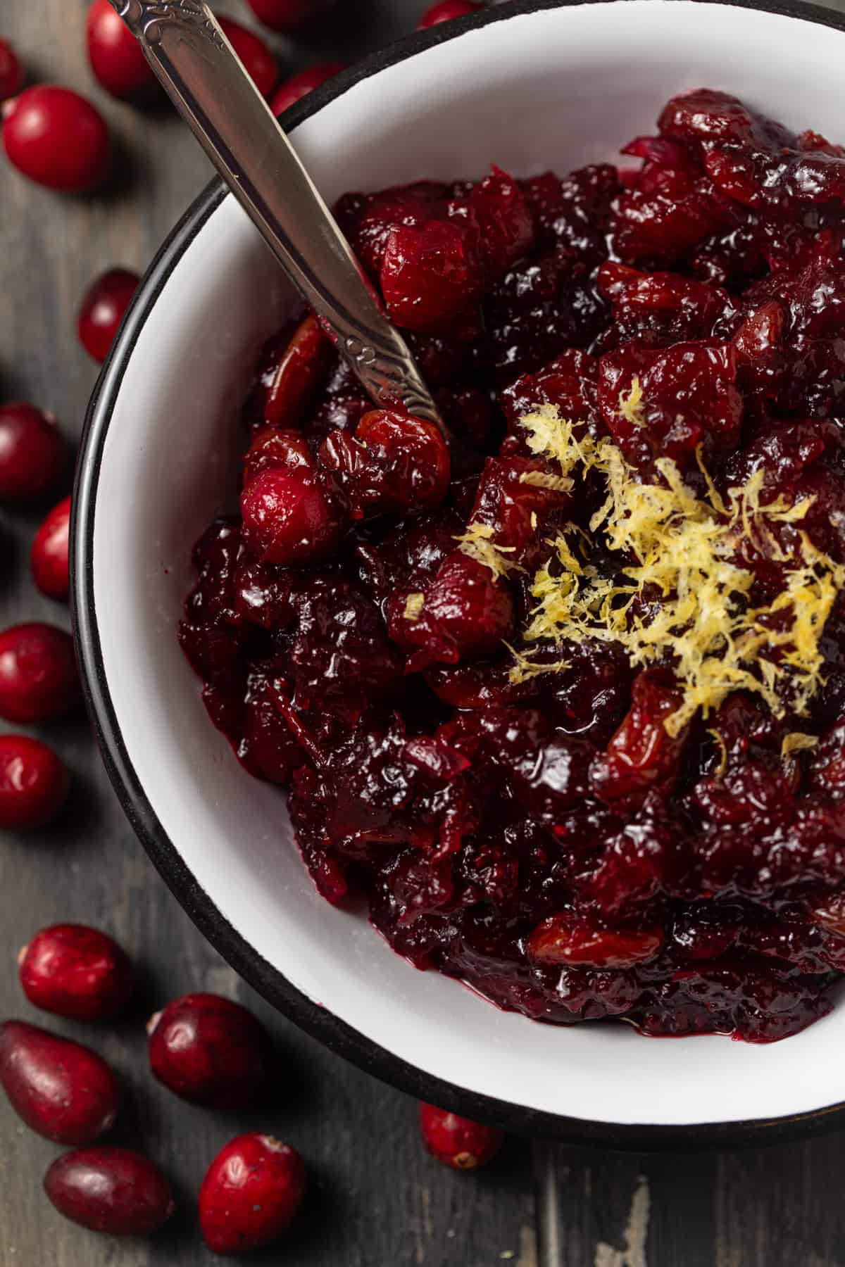 Cranberry sauce topped with lemon zest in a white bowl with a silver spoon. 