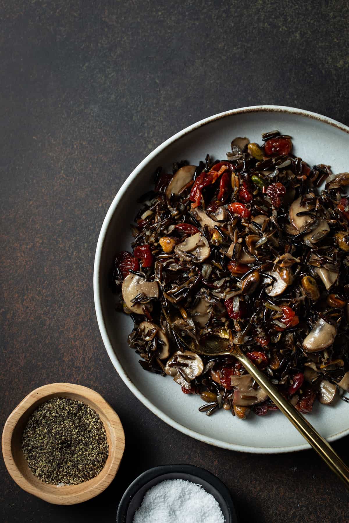 Gray bowl filled with cooked wild rice, pistachios, mushrooms & cherries next to two small bowls of salt & pepper. 