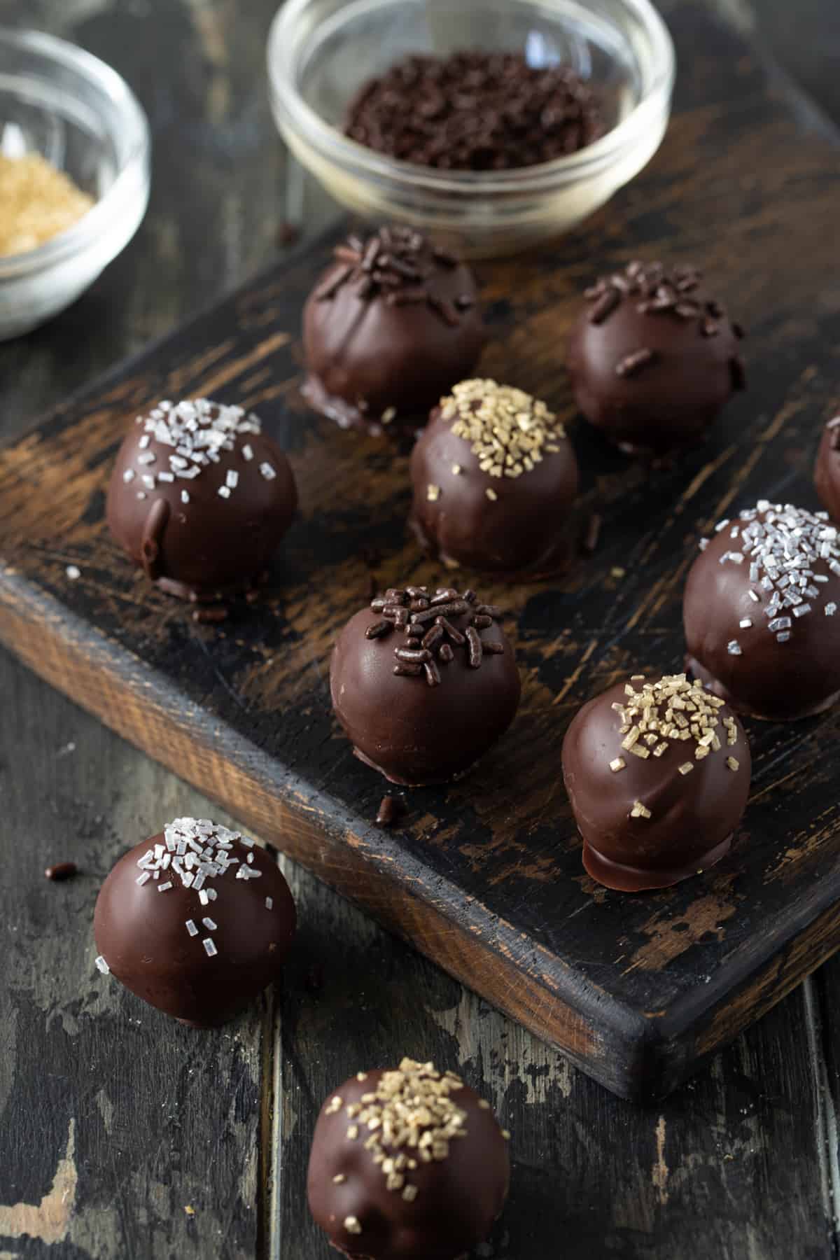 Dipped truffles on a wooden board. Small glass bowls of sprinkles in the background. 