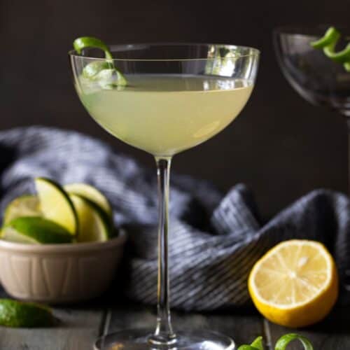tall martini glass filled with drink on table with lemon wedges.