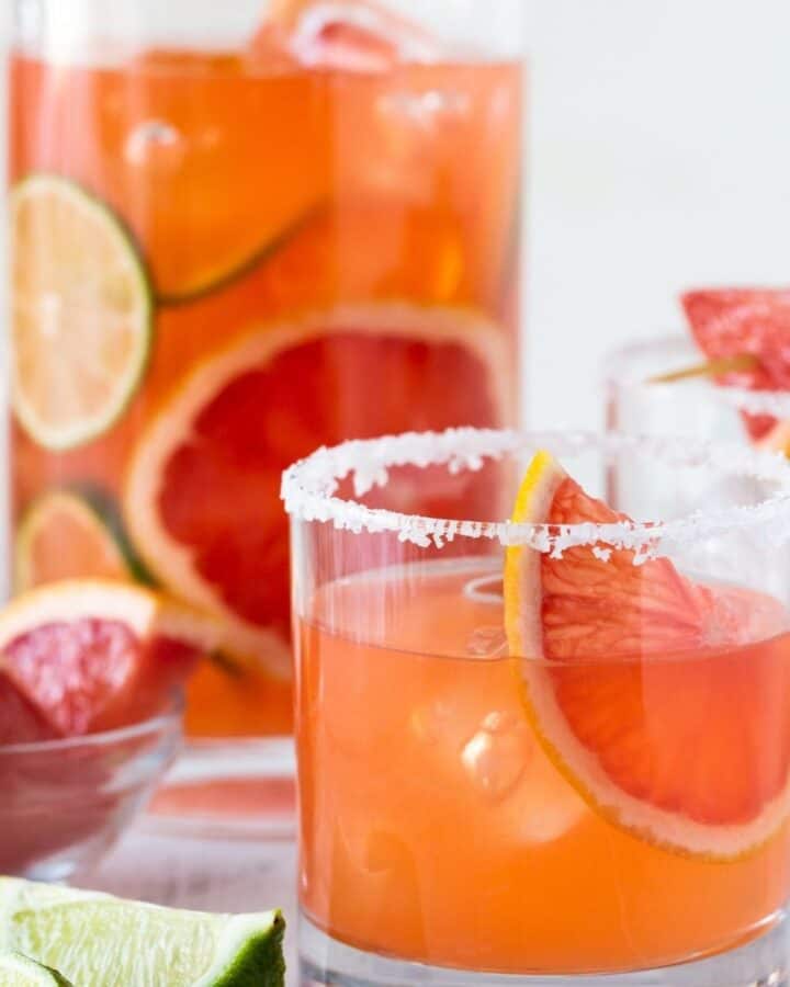 low ball glass with salted rim filled with drink and grapefruit wedge with pitcher behind it.