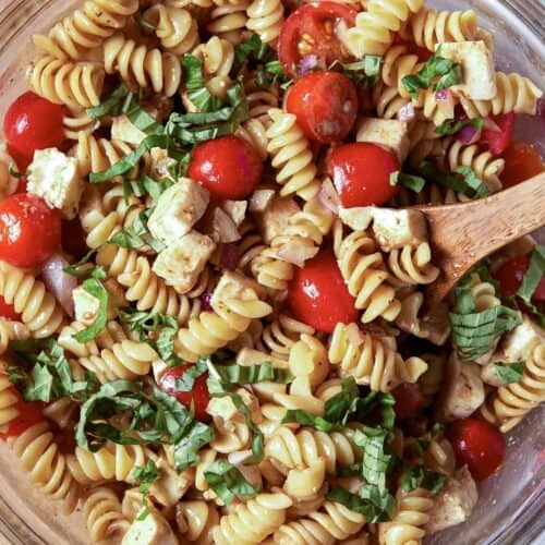 Caprese pasta salad in a glass bowl with a wooden spoon on the side.