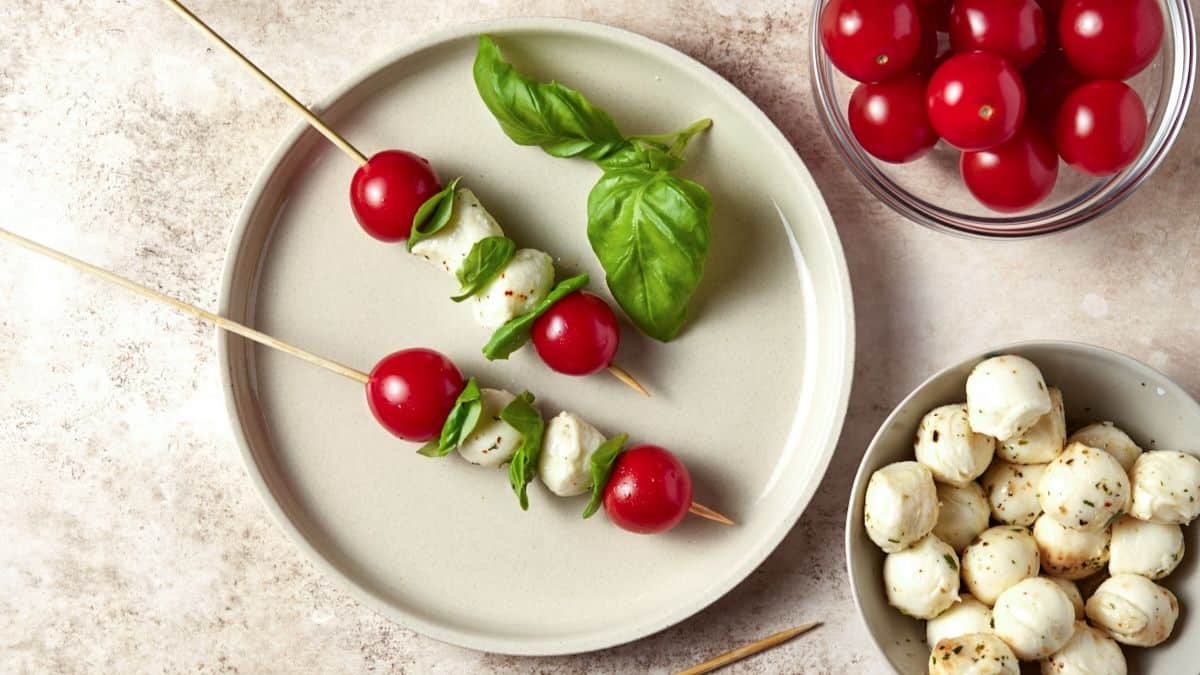 Two Caprese Skewers on a light plate with bowls of tomatoes and mozzarella balls nearby. 