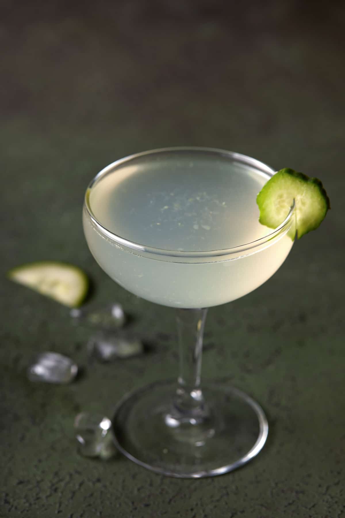 Vodka gimlet in a coupe glass garnished with cucumber on a dark green board.