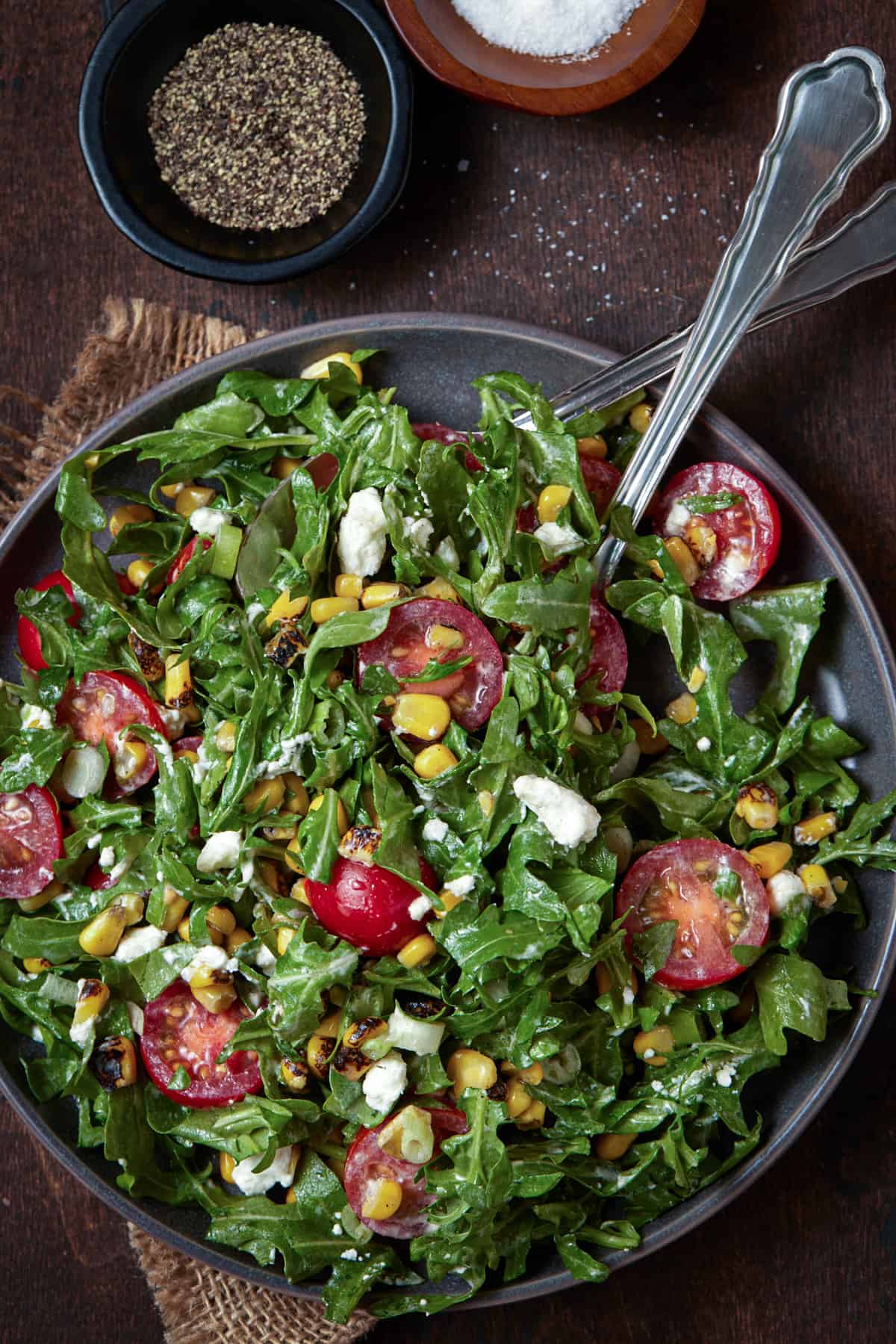 Plate of arugula salad with tomatoes, salt and pepper and serving spoons.