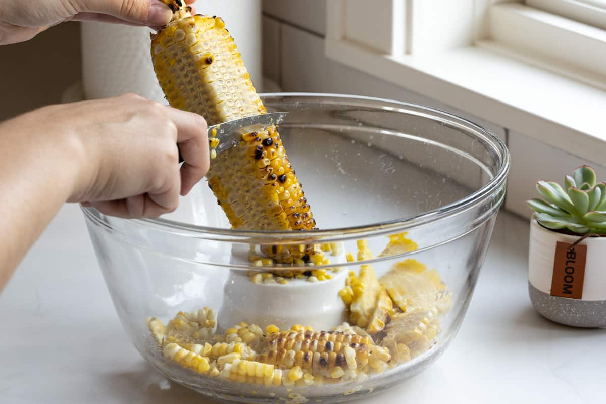Hand cutting grilled corn on the cob into a glass bowl. 