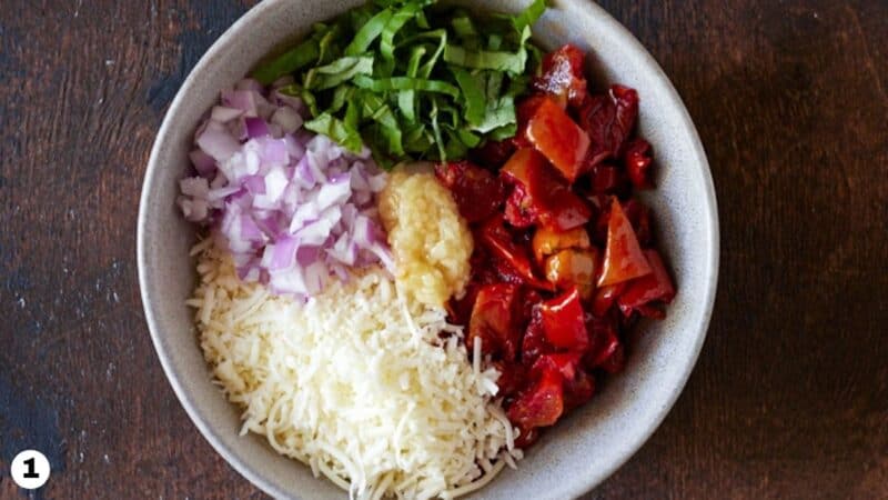 Chopped roasted tomatoes, shredded asiago cheese, chopped onions, basil and garlic in a white bowl.