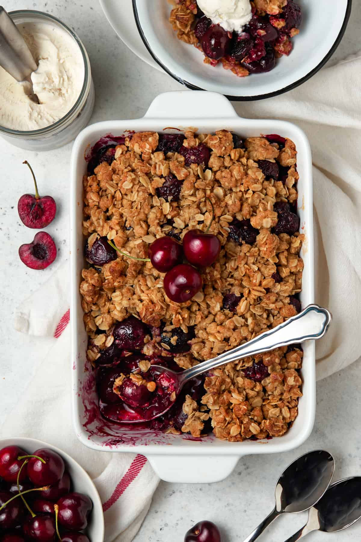 White baking dish of Cherry Crisp with serving removed. Bowl of fresh cherries and vanilla ice cream nearby. 