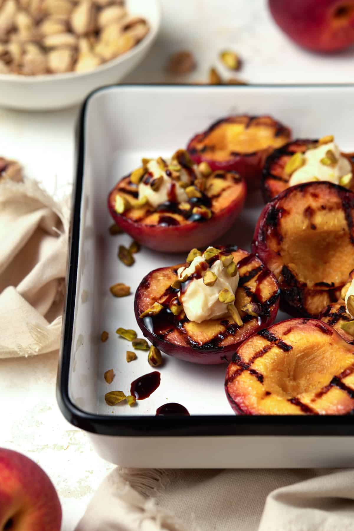 Grilled peach halves in a white enamel tray drizzled with balsamic glaze. 