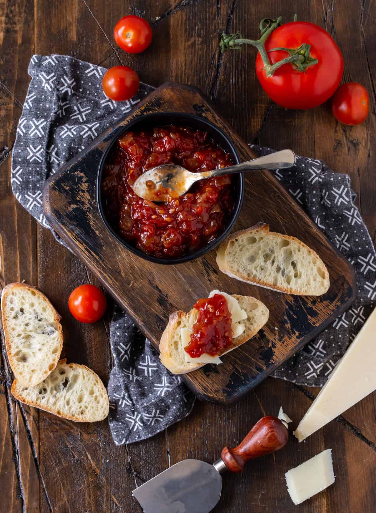 Baguette and cheese slices topped with Sweet and Tangy Tomato Jam recipe on a wooden cutting board.