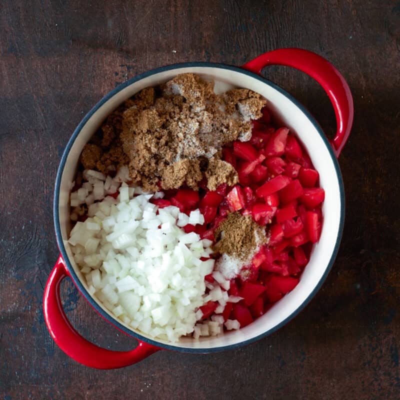 Ingredients for tomato jam in a red dutch oven, including chopped tomatoes, onion, spices and sugar.
