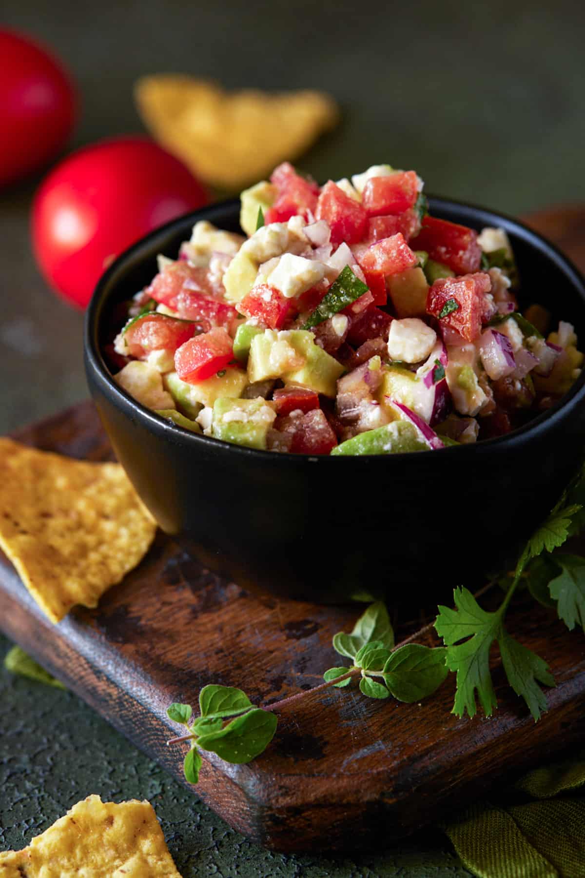 Bowl of avocado salsa wiht feta cheese on table with chips and fresh herbs.