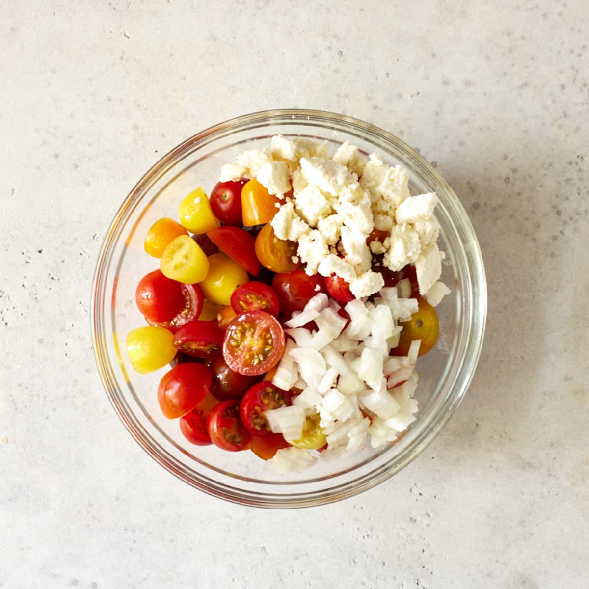 Halved cherry tomatoes, chopped onion and feta cheese in a glass bowl.