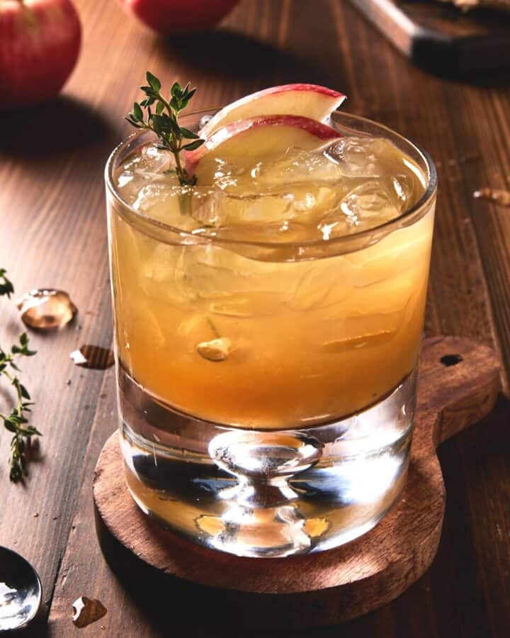 Apple cider and bourbon cocktail in lowball glass with fresh apple slices and thyme sprigs.