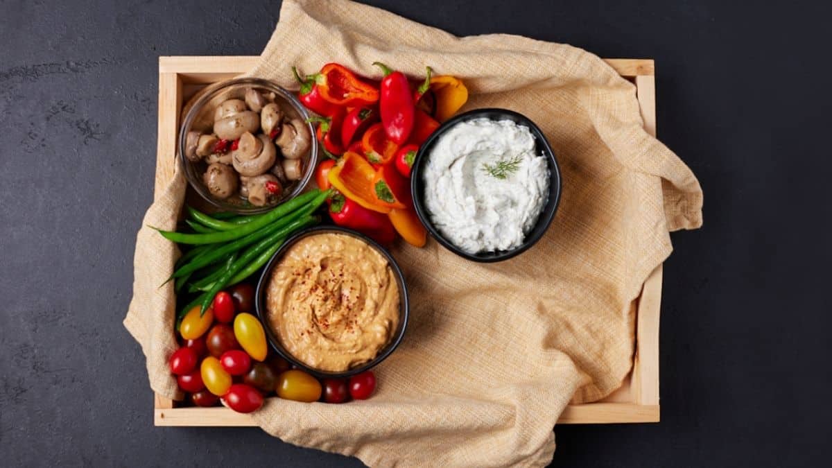 Cut up veggies on a wooden platter with bowls of dip. 