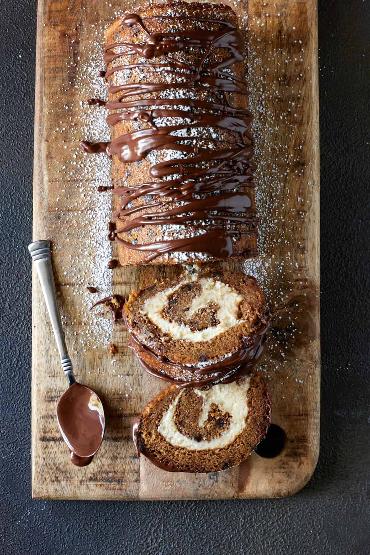 Pumpkin Chocolate Swiss Roll Cake recipe on a wooden board with a spoon dipped in chocolate. 