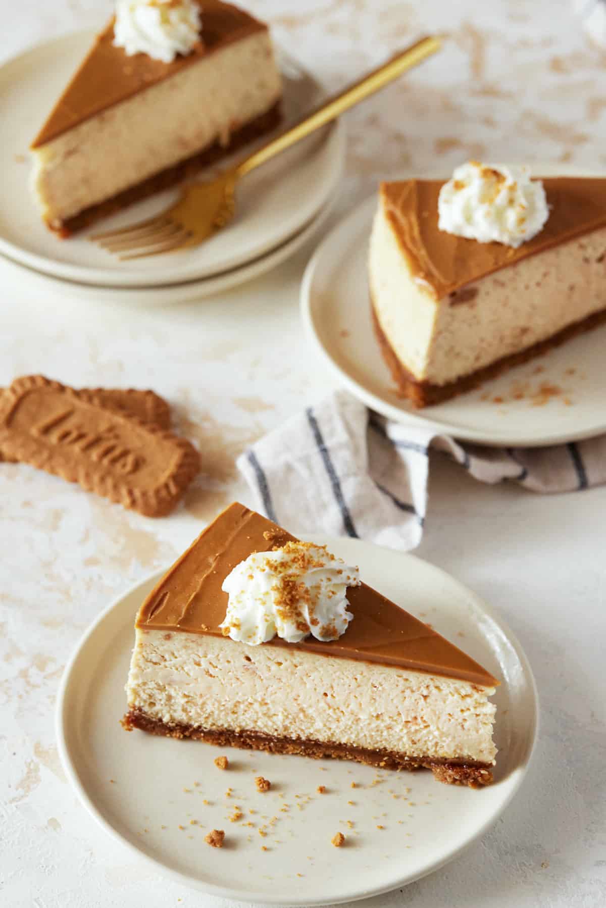 Slices of Biscoff Cheesecake on cream plates. Topped with whipped cream and cookie crumbs. 