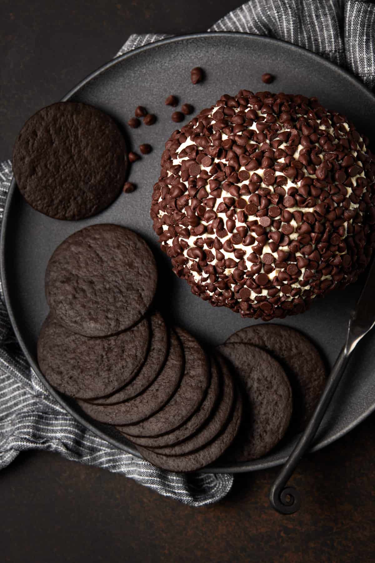 Cheesecake ball with chocolate cookies and spreader on gray plate.