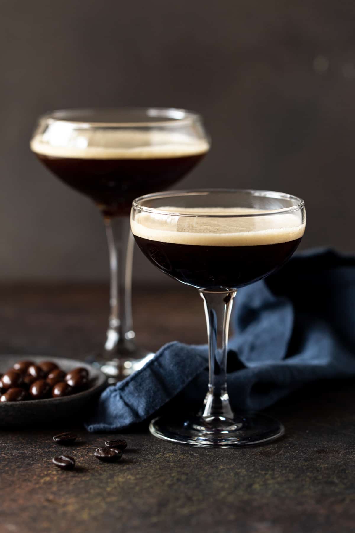Two espresso martinis in glasses on a brown board with navy napkin. 