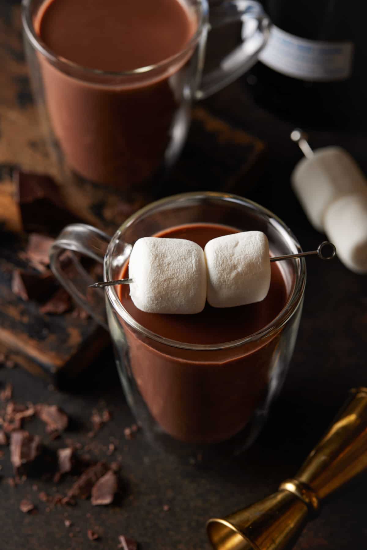 A glass mug of hot chocolate with two large marshmallows on a skewer. 