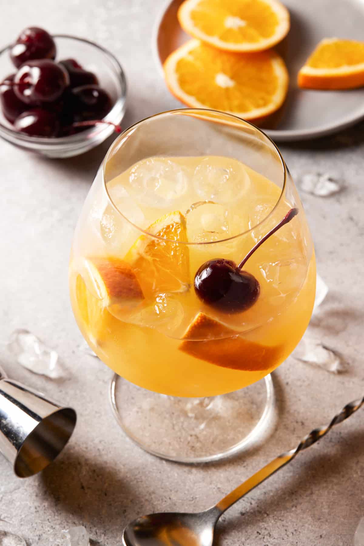 Orange juice cocktail in a footed glass filled with ice and garnished with cocktail cherries and orange slices. 