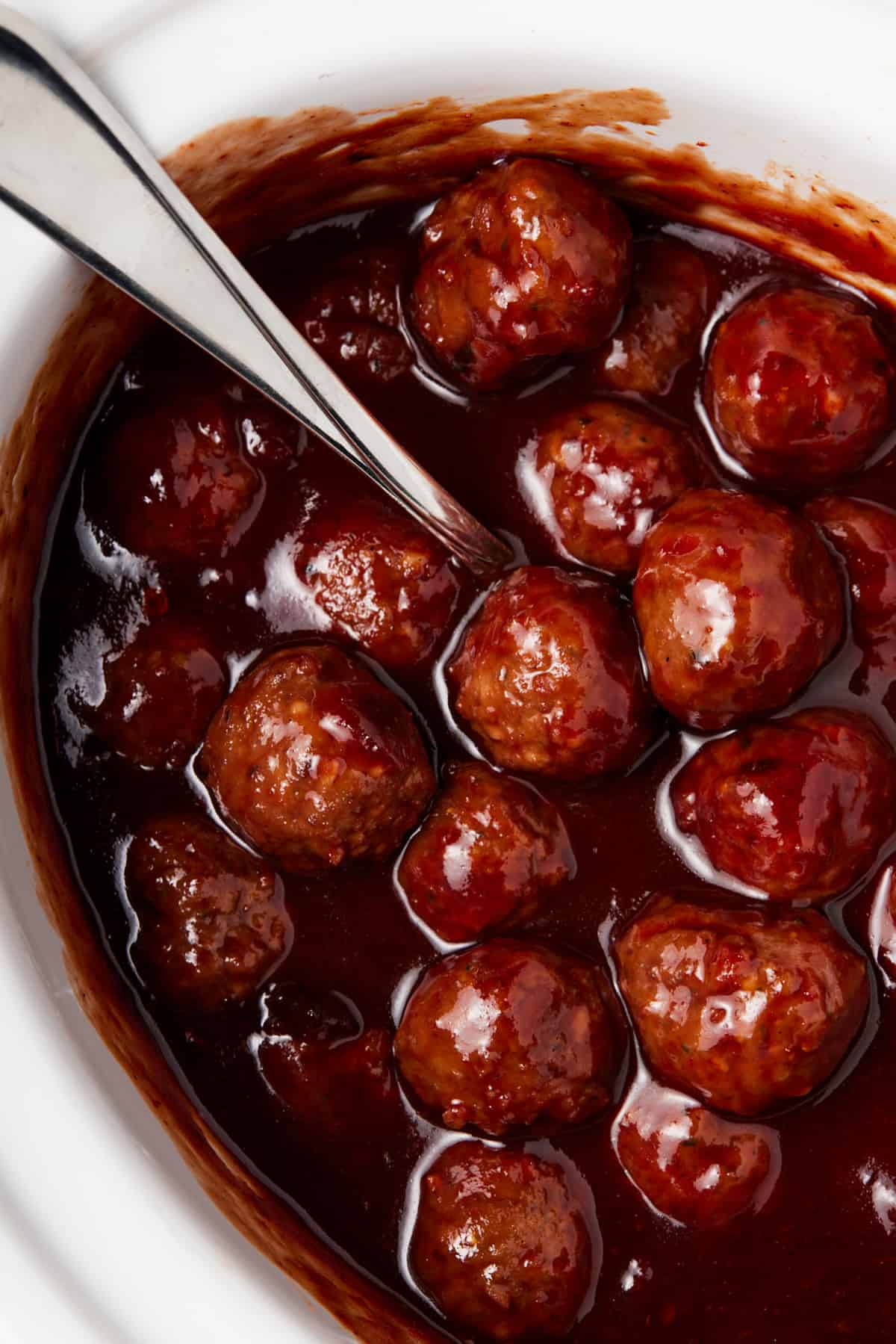 White slow cooker filled with bbq meatballs with spoon.