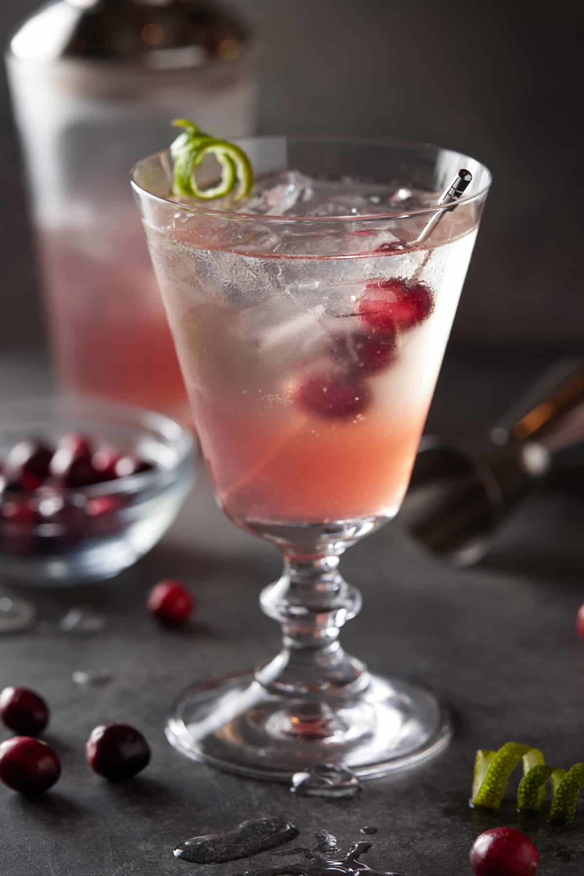 A footed glass filled with cranberry gin cocktail and garnished with fresh cranberries and a lime twist. 