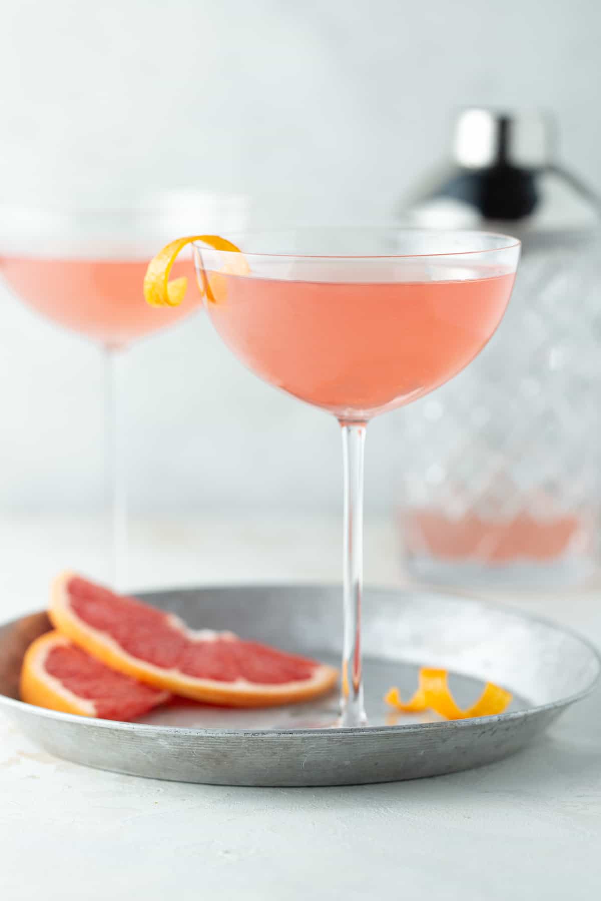 2 pink martinis with a shaker in the background on a white surface.
