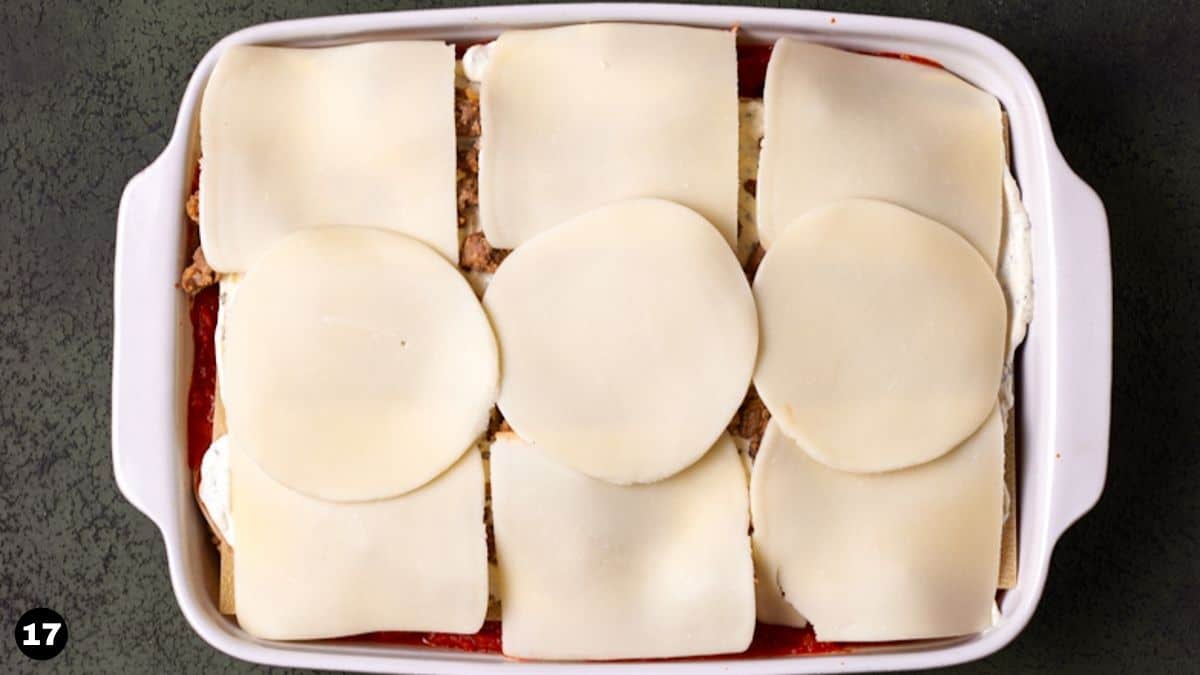Cheese slices on top of unbaked layered lasagna in white casserole dish. 
