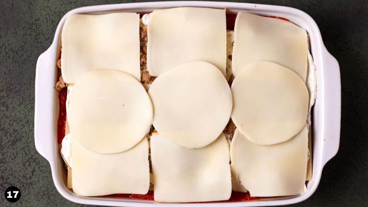 Cheese slices on top of unbaked layered lasagna in white casserole dish.
