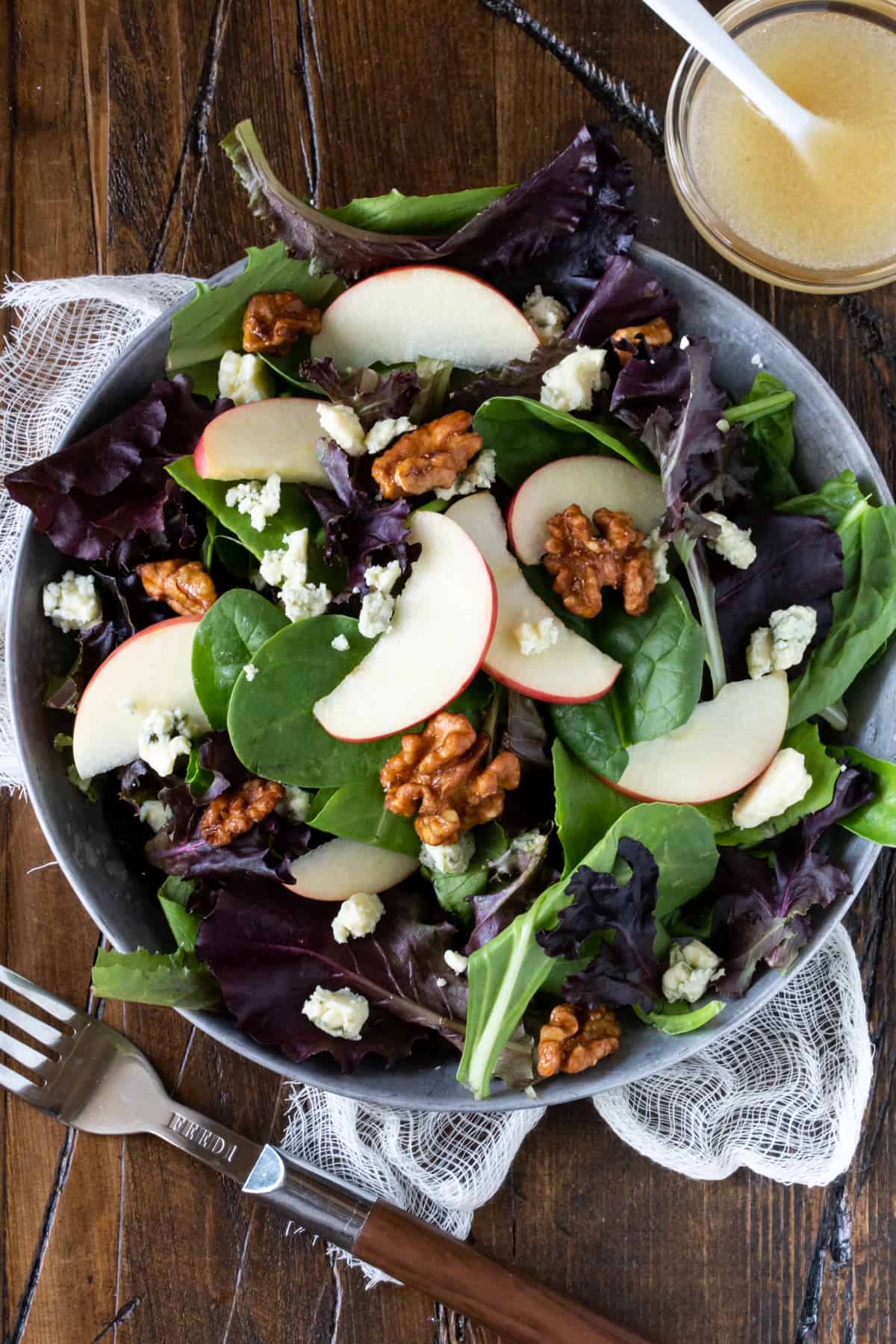 Silver bowl of Mixed Green Salad with Blue Cheese Crumbles, Apples and Candied Walnuts. Next to salad is a small glass bowl of salad dressing. 