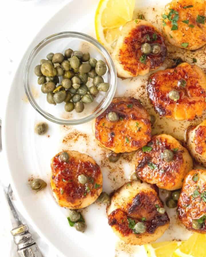 White plate with seared scallops, capers and lemon slices.