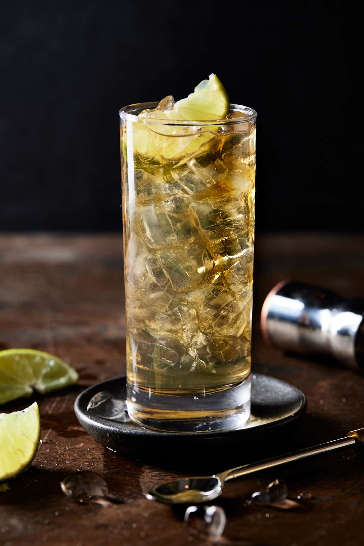 Highball glass of whiskey ginger drink with a lime wedge on a black coaster. 