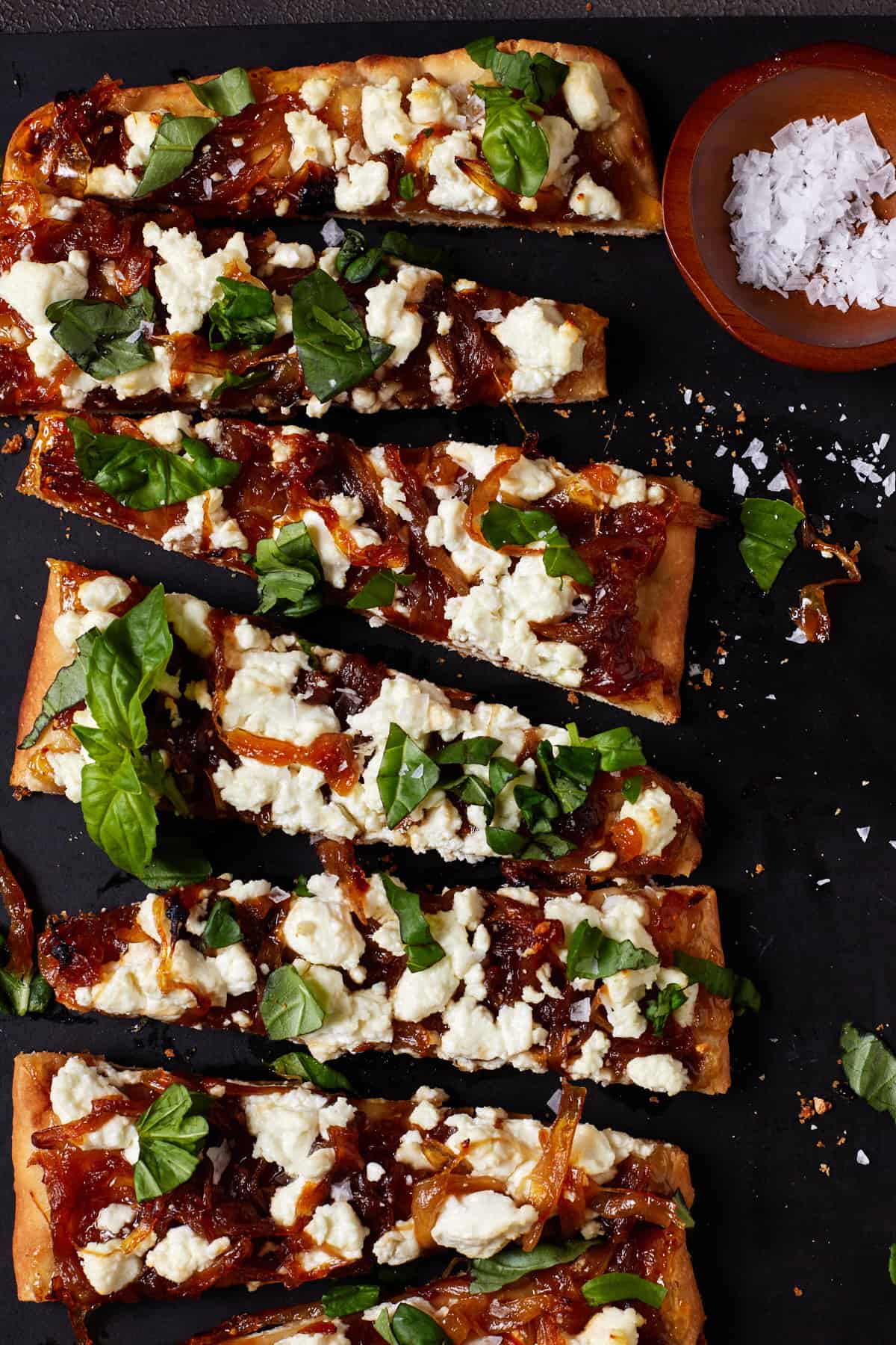 Slices of Goat Cheese, Onion & Basil pizza on a black cutting board with a small bowl of salt. 