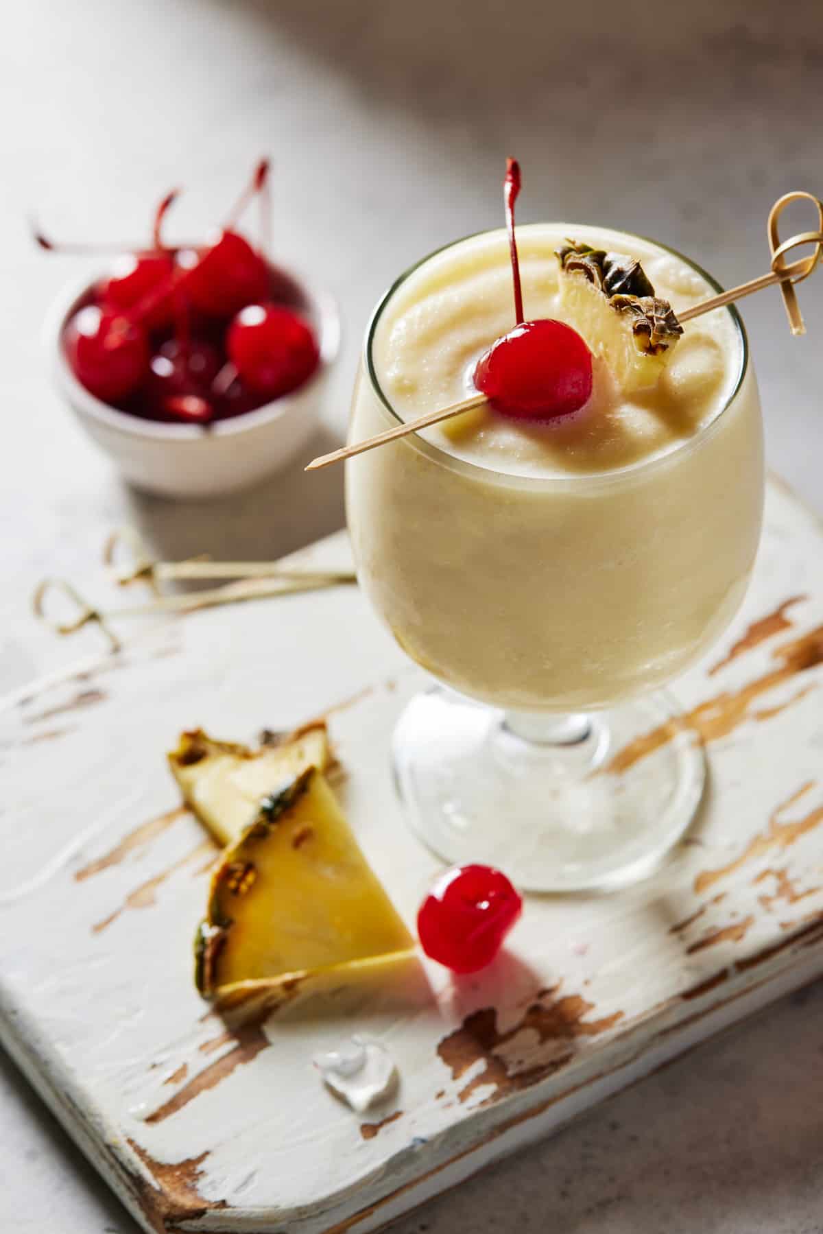 Footed glass of virgin piña colada with small bowl of maraschino cherries and pineapple wedges. 