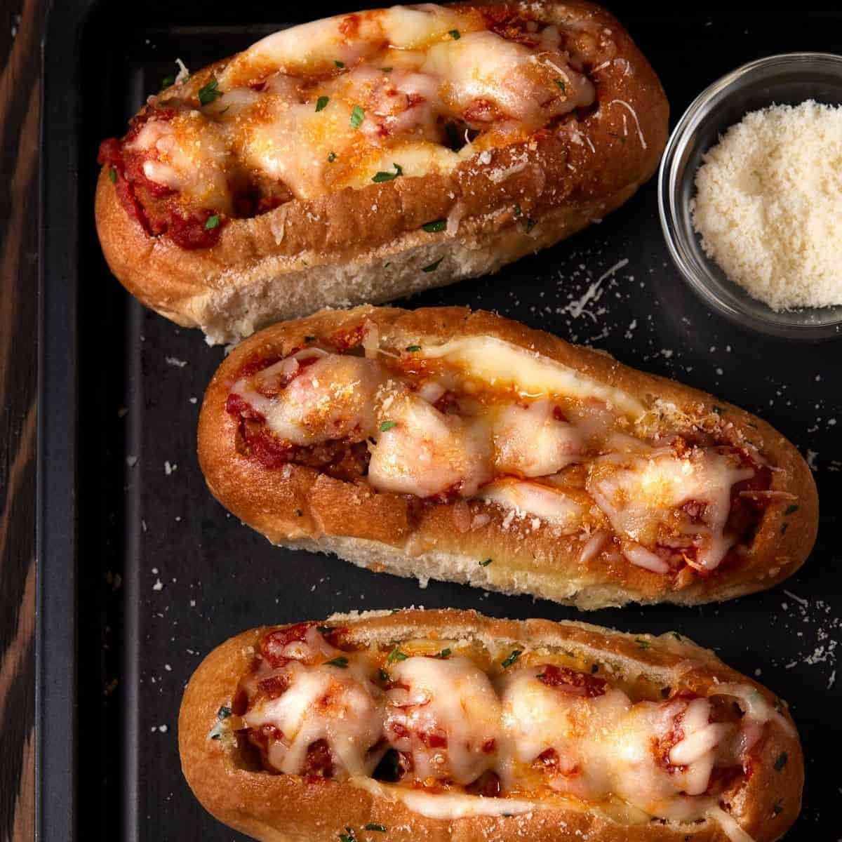 Toasted meatball subs on a dark sheet pan with small bowl of grated parmesan cheese. 