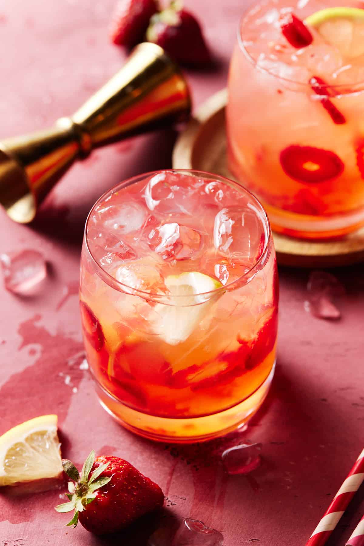 Two low ball glasses filled with strawberry vodka lemonade on a pink board. 