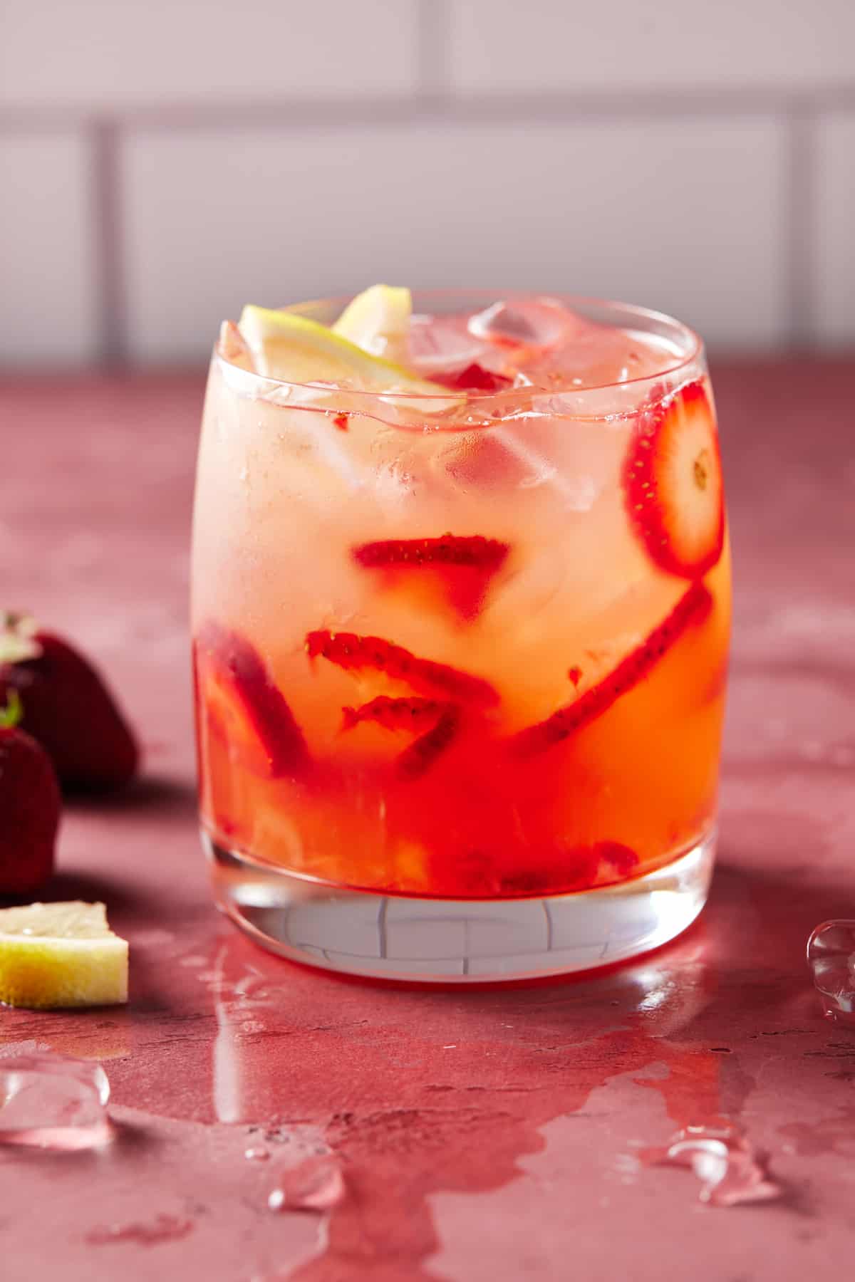 Lowball glass filled with strawberry vodka lemonade, strawberry slices and ice on a pink board.