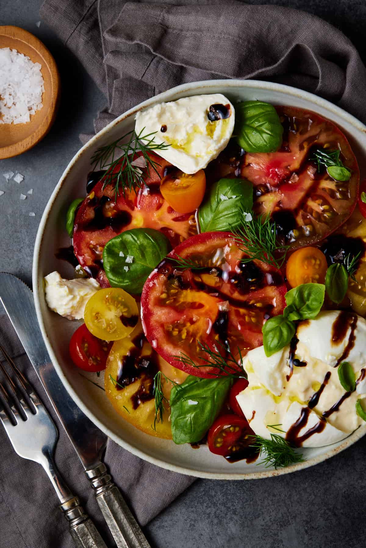 Light grey bowl with burrata caprese salad, a knife and fork. 