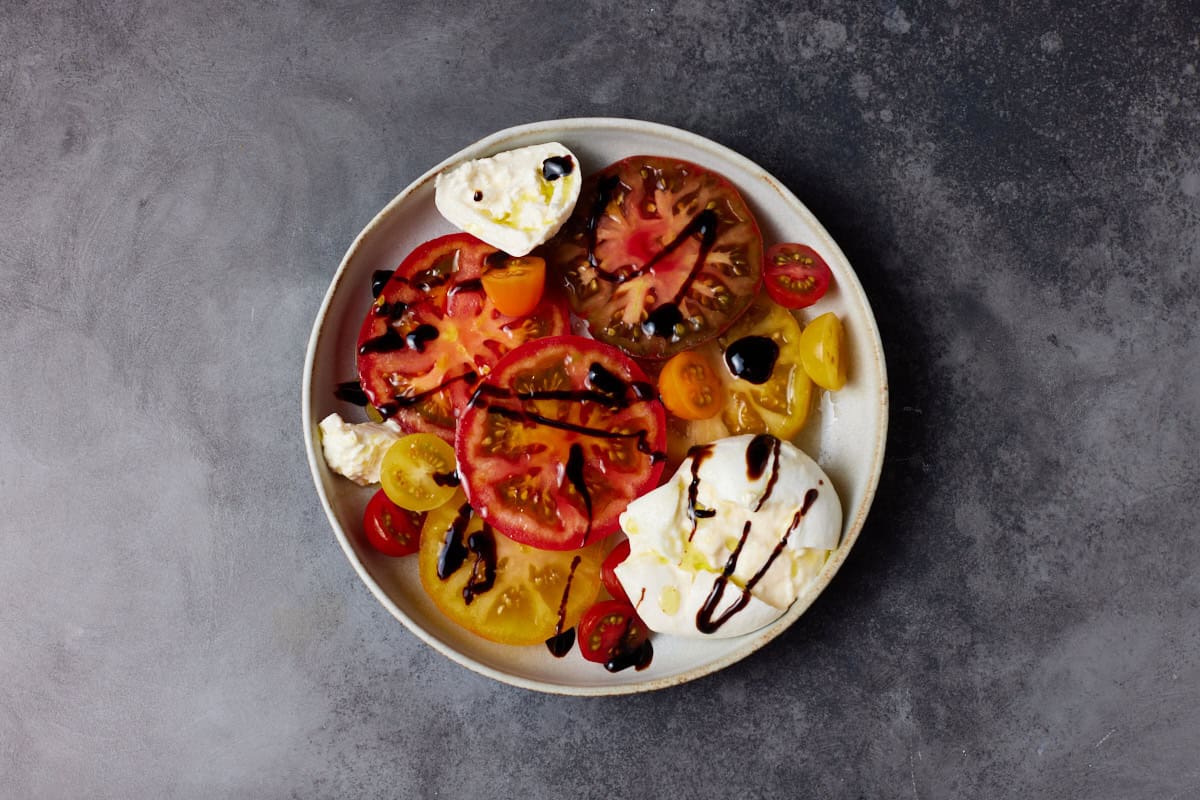 Sliced heirloom tomatoes, burrata cheese  and balsamic glaze in a shallow bowl. 