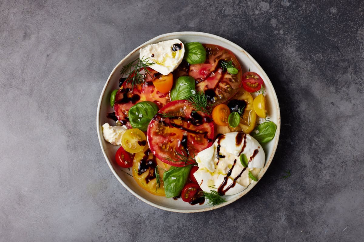 Sliced heirloom tomatoes burrata cheese, balsamic glaze and fresh herbs in a shallow bowl. 